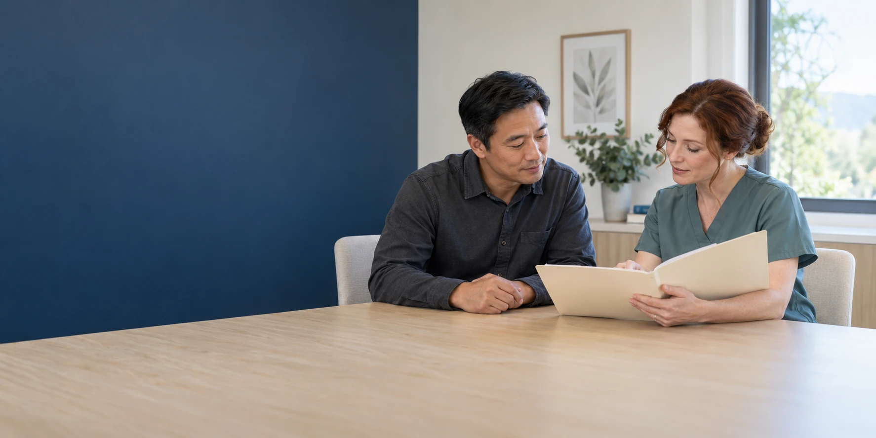 An adult patient in a work shirt and a clinician discussing an open folder at a consultation desk.